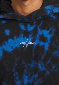 Černá mikina s kapucí s modrým tie-dye vzorem, zdobená bílým nápisem. Materiál vypadá měkký, má standardní střih a nastavitelnou kapuci.