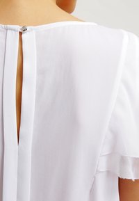 Bílá blůza s klíčovou dírou na zádech, zapínání na stříbrný knoflík, měkká tkanina, krátké volánové rukávy a mírná textura.