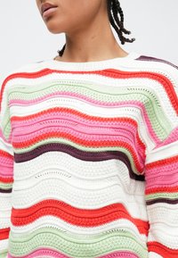 Άτομο που φοράει πολύχρωμο πλεκτό πουλόβερ με κυματιστές ρίγες σε λευκό, κόκκινο, ροζ, πράσινο και μοβ, φαίνεται από τον λαιμό μέχρι τη μέση.