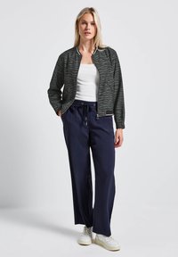 Veste bomber à motif zèbre en vert foncé et noir, associée à un pantalon bleu marine à coupe ample et des baskets blanches. Texture lisse et design décontracté.