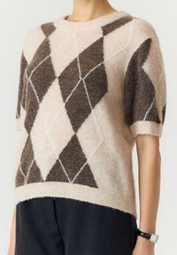 Personne portant un pull beige doux à manches courtes avec un grand motif losange marron et blanc et un pantalon foncé, montrant le milieu du torse et le bras.