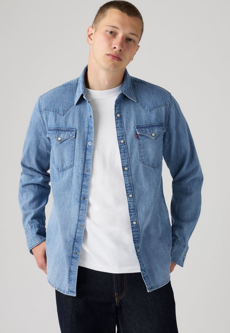 Jeune homme portant une chemise en jean bleu clair par-dessus un t-shirt blanc et un pantalon foncé, debout devant un fond gris clair.