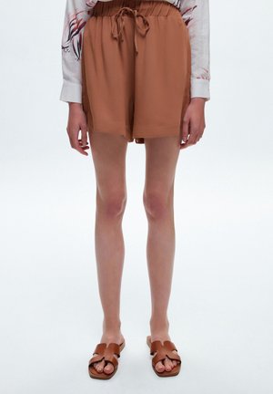 Persoon die lichtbruine shorts met trekkoord draagt, een wit overhemd met lange mouwen met rode patronen, en bruine slippers, staand op een witte achtergrond.