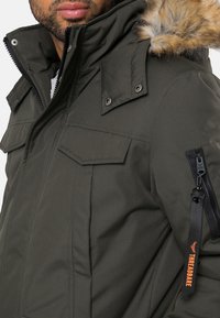 Hombre vistiendo una chaqueta de invierno verde oscuro con capucha forrada de pelaje, bolsillos en el pecho y un bolsillo con cremallera en la manga con una etiqueta negra y naranja de "THREADBARE".
