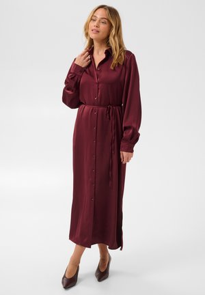 Femme portant une robe bordeaux à manches longues, boutonnée, avec une ceinture à la taille et des chaussures foncées à bout pointu, debout devant un fond uni.