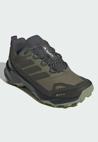 GORE-TEX planinarska cipela u tamnozelenoj i sivoj boji. Ima mrežasti gornji deo, gumeni đon i teksturirane detalje, s tri crne pruge sa strane.