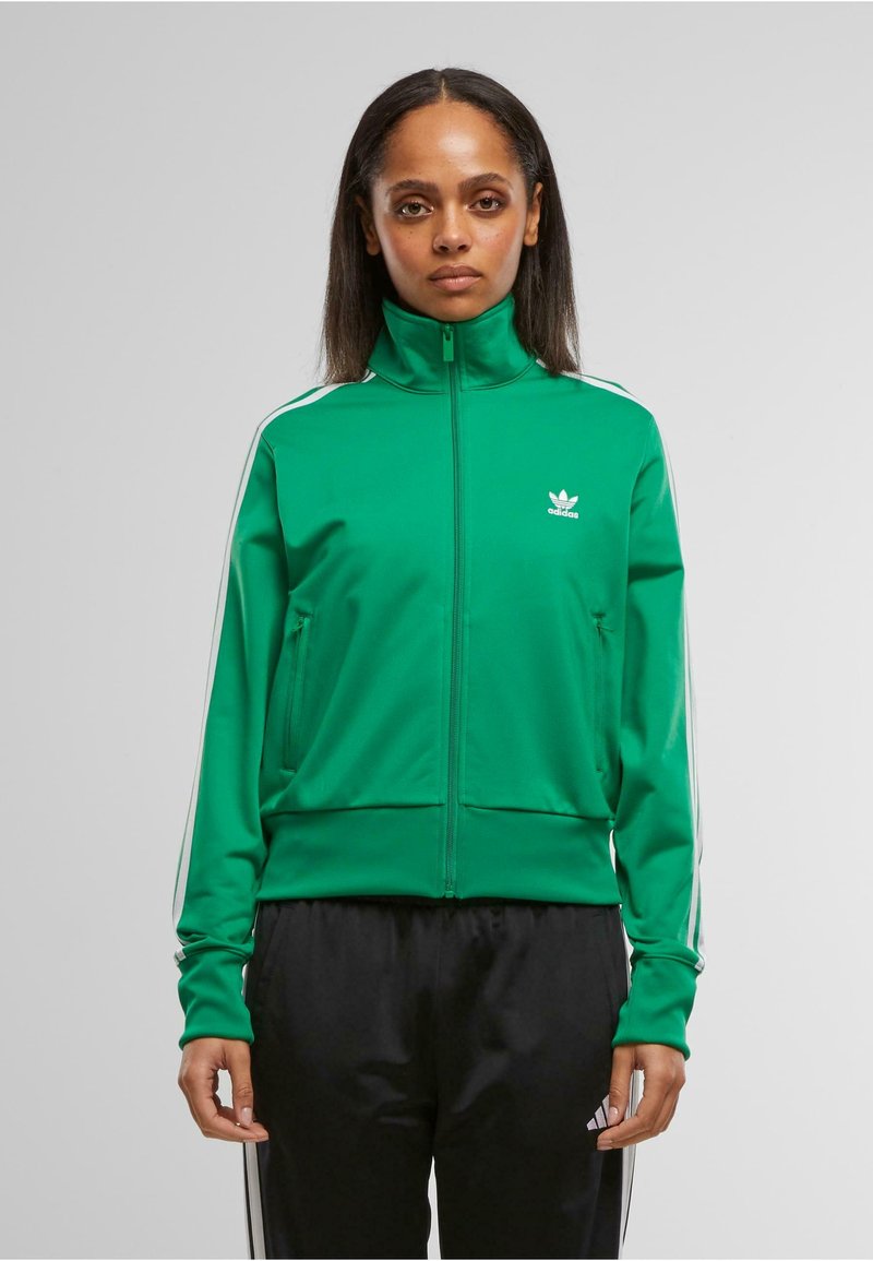 Chaqueta deportiva verde con cremallera y cuello alto, acentos de tres franjas blancas en las mangas y logo bordado en el pecho. Tejido suave y liso.