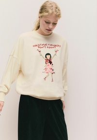 Kremowy sweatshirt o luźnym kroju, z grafiką dziewczyny w różowej sukience trzymającej koktajle i napisem: "Kalorie na Boże Narodzenie się nie liczą."
