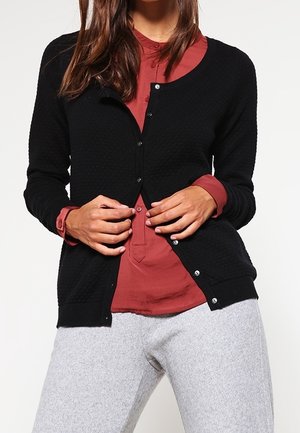 Frau, die eine schwarze Strickjacke über einem rostfarbenen Hemd zuknöpft, trägt hellgraue Hose und steht vor einem weißen Hintergrund.