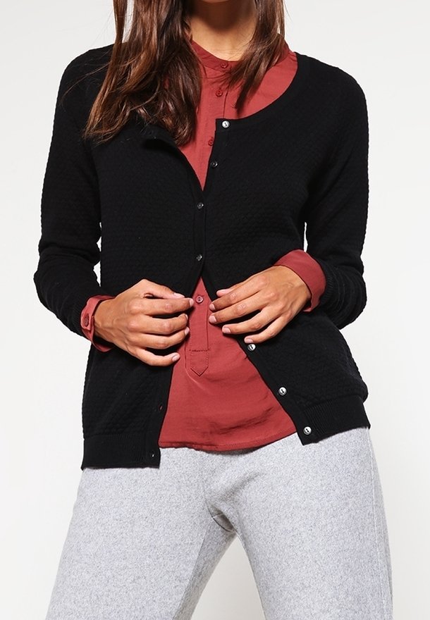 Femme boutonnant un cardigan noir sur une chemise couleur rouille, portant un pantalon gris clair, debout devant un fond blanc.