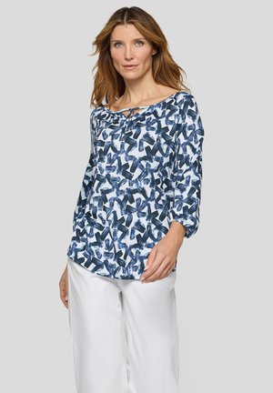 Woman with light brown hair wears a white blouse with dark blue abstract brushstroke pattern and white pants against a gray background.