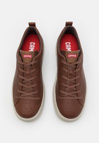 Brown leather sneakers with a smooth texture, featuring a red insole, brown laces, perforated detailing on the toe, and a white sole.