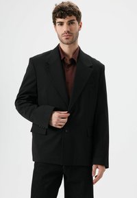 Zwarte blazer met een gestructureerd design, een enkele knoopsluiting en twee voorzakken. Gecombineerd met een donkerrood overhemd eronder.