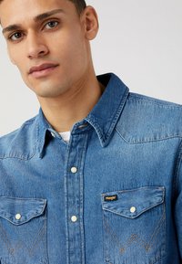 Camisa de mezclilla azul con cuello puntiagudo, bolsillos frontales y cierres de botones blancos. Presenta costuras en contraste y un logo de Wrangler en el bolsillo del pecho.