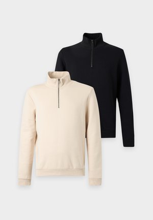 Jeune homme aux cheveux bouclés portant un pull beige à fermeture éclair, un jean bleu ample et des chaussures décontractées marron, debout les mains dans les poches.