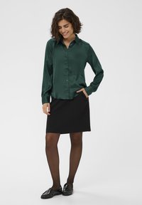 Green satin blouse with a collar, button-down front, and long puff sleeves paired with a black knee-length skirt. Black loafers and tights.