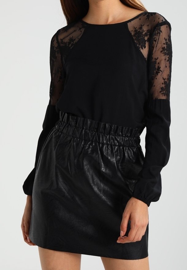 Femme portant une robe noire avec des manches longues en dentelle florale transparente et une jupe en faux cuir à taille froncée.