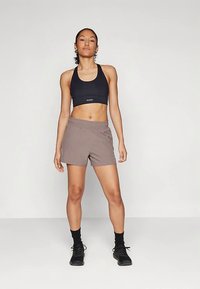 Naine seisab enesekindlalt mustas spordibrassis, taupe värvi spordilühikeses seelikus, mustade sokkide ja mustade tossudega lihtsa tausta ees.