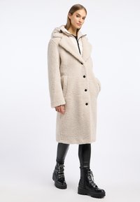 Beige, textuur, oversized jas met een capuchon, dubbele sluiting, zwarte knopen en zijzakken. Draagt over zwarte leggings en laarzen.