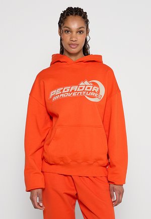 Junge Frau mit geflochtenem Haar, die einen orangefarbenen Hoodie und passende Hose mit dem Logo "Pegador Adventures" auf der Vorderseite trägt.