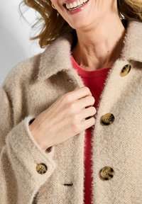 Beige wollen mix jas met een gestructureerde afwerking, grote bruine knopen en een notch-kraag, gelaagd over een rode trui.