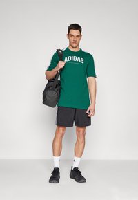 Zöld Adidas póló fehér logóval, fekete rövidnadrág és fekete táska. A modellen fekete sportcipők és fehér zoknik vannak, háttérben szürke fal.