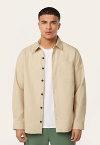 Beige overhemd met knopen en een kraag, met een borstzakje en zwarte knopen. Gedragen over een wit T-shirt, gecombineerd met groene broek.