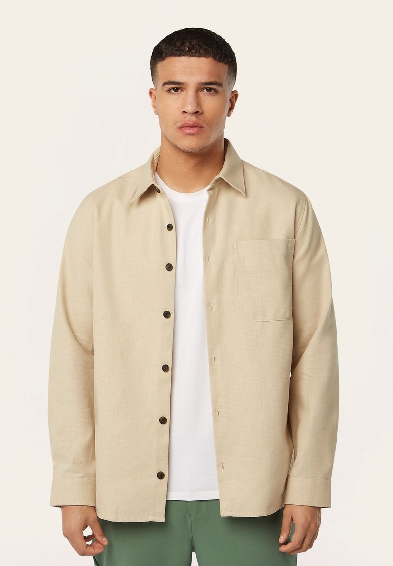 Beige overhemd met knopen en een kraag, met een borstzakje en zwarte knopen. Gedragen over een wit T-shirt, gecombineerd met groene broek.