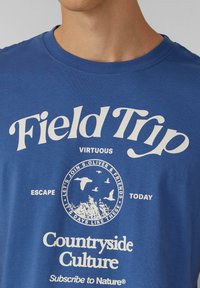 Niebieska bawełniana koszulka z krótkim rękawem z białym nadrukiem tekstu i grafik, w tym z hasłem "Wycieczka" oraz motywami przyrodniczymi, z okrągłym dekoltem.