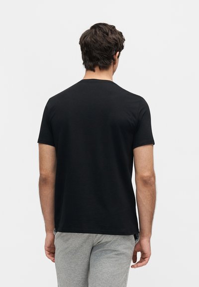 Άνδρας με σκούρα μαλλιά που φοράει απλό μαύρο κοντομάνικο μπλουζάκι και ανοιχτό γκρι παντελόνι, βλέπεται από πίσω με φόντο ένα λευκό φόντο.