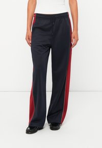 Marineblauwe trainingsbroek met rode zijstrepen, een elastische tailleband en een trekkoord. De stof lijkt glad, met een subtiele glans en een losse pasvorm.