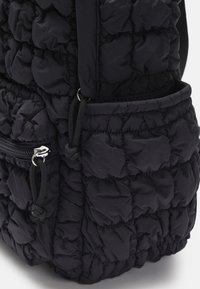 Mochila negra acolchada de tela suave. Presenta un bolsillo con cremallera y un bolsillo lateral de malla, con cierre de cordón y superficie texturizada.