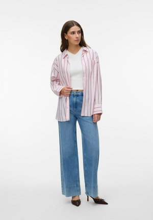 Donna che indossa un crop top bianco, una camicia rosa a righe oversize, jeans blu a gamba larga e scarpe marroni con tacco a punta, in piedi su sfondo bianco.
