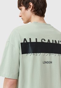 Camiseta extra grande de color verde claro con texto negro, elementos gráficos en la parte posterior y un escote relajado. Fabricada en algodón y con mangas cortas.
