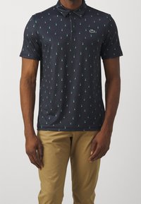 Man wearing navy blue polo shirt with small green and white golfer icons and khaki pants, standing against plain background.