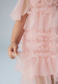 Rožnata obleka iz tila z več plastmi volanov, prosojnimi rokavi in teksturiranim dizajnom. Roka drži spodnji del obleke.