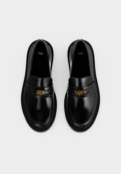 Paire de mocassins en cuir noir avec un détail tête de lion doré sur les brides, marqués "maje" à l'intérieur, vus de dessus sur fond blanc.