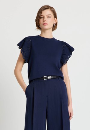 Giovane donna che indossa una maglia blu navy con maniche a volant in pizzo traforato e pantaloni a vita alta coordinati, in piedi con una mano in tasca.