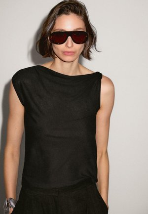 Un haut sans manches noir avec un col bateau, fabriqué à partir d'un tissu léger ; le modèle porte des lunettes de soleil sombres et a les cheveux courts et texturés.