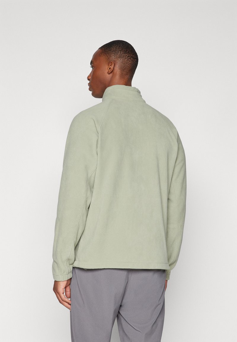 Chaqueta de forro polar verde claro con un collar alto, costuras laterales y puños elásticos, llevada sobre pantalones grises, vista desde la parte trasera.