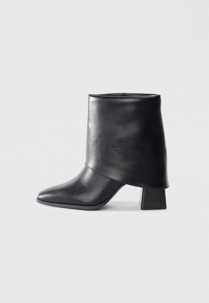 Bottine en cuir noir avec bout pointu et talon carré sur un fond gris clair uni.