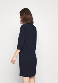 Marineblå klänning med trekvartslånga ärmar, rak skärning och sidfickor. Tyget verkar slätt med minimal detaljering.