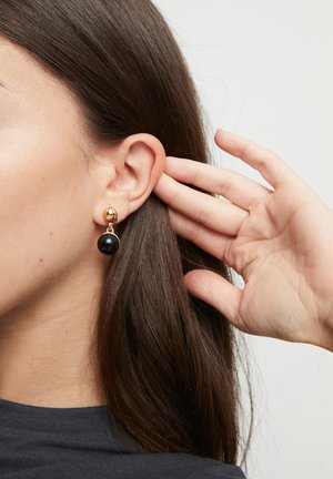 Mujer acomodándose el cabello largo y castaño detrás de la oreja, lleva pendientes de botón dorados con gotas esféricas negras, contra un fondo claro y liso.