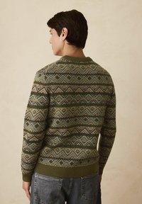 Egy férfi háttal áll, olívazöld és bézs geometrikus mintájú pulóvert visel kék farmernadrággal, sima bézs háttér előtt.