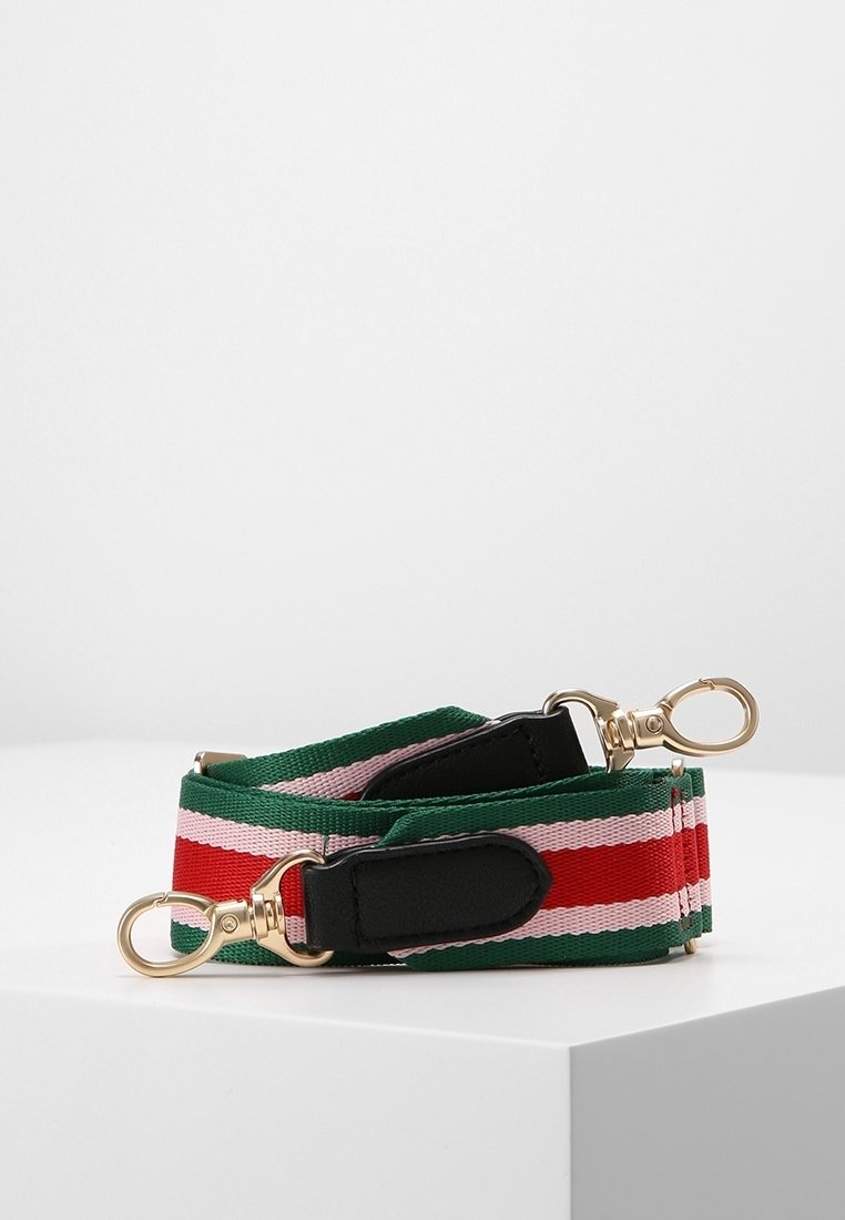 Sangle de sac en tissu rayée verte, rouge et rose avec des fermoirs pivotants dorés et des accents en cuir noir, enroulée sur une surface blanche.