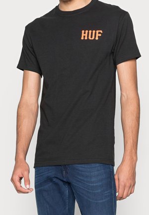 Sort bomulds t-shirt med rund halsudskæring og korte ærmer. Den har et orange "HUF" logo på venstre bryst. Båret med blå jeans.