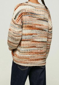 Personne portant un pull en maille épaisse avec des rayures multicolores horizontales en beige, orange, gris et marron, associé à un jean bleu foncé.