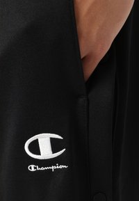 Čierna tkanina má výrazné biele vyšívané logo Champion. Dizajn obsahuje vrecko a detail gombíka pre funkčnosť.