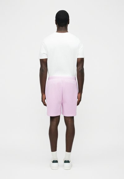 Mand stående med ryggen mod, iført en hvid kortærmet skjorte, lyserøde shorts, hvide sokker og hvide sneakers med mørkegrønne detaljer.