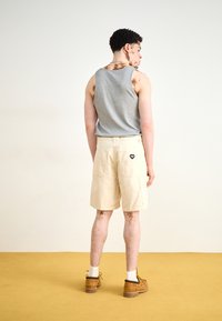 Grå ermeløs tanktop, beige shorts med baklommer og brune sko. Modellen vender bort, noe som avslører et hjerte-logo på shortsene.